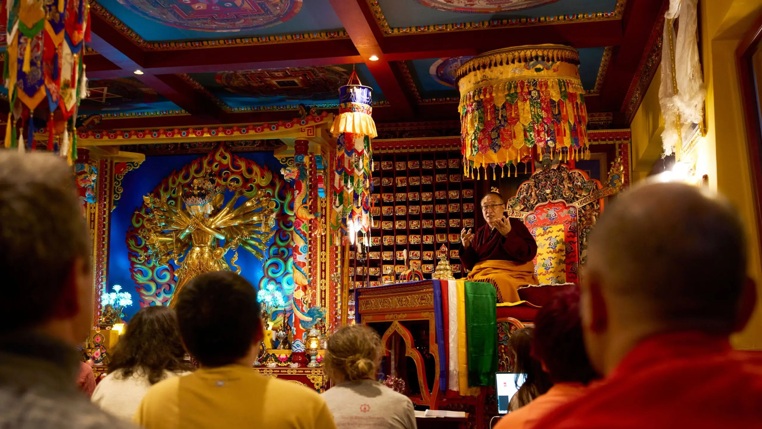 Khentrul Rinpoche sitting on the throne teaching in the Kalachakra temple, The Kalachakra statue in the background is lit with glowing blue lights. 